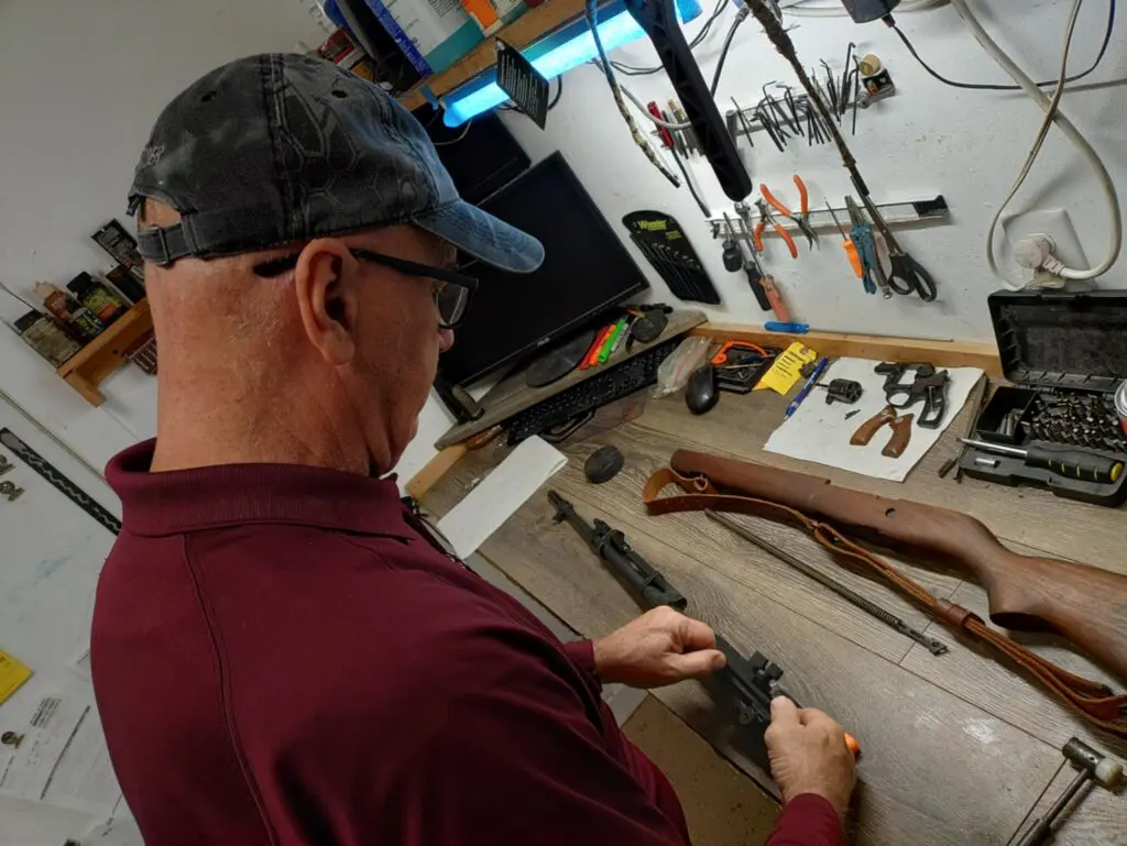 An older man in a maroon shirt and baseball cap is leaning over a wooden workbench, working on a disassembled firearm, surrounded by tools and gun parts.