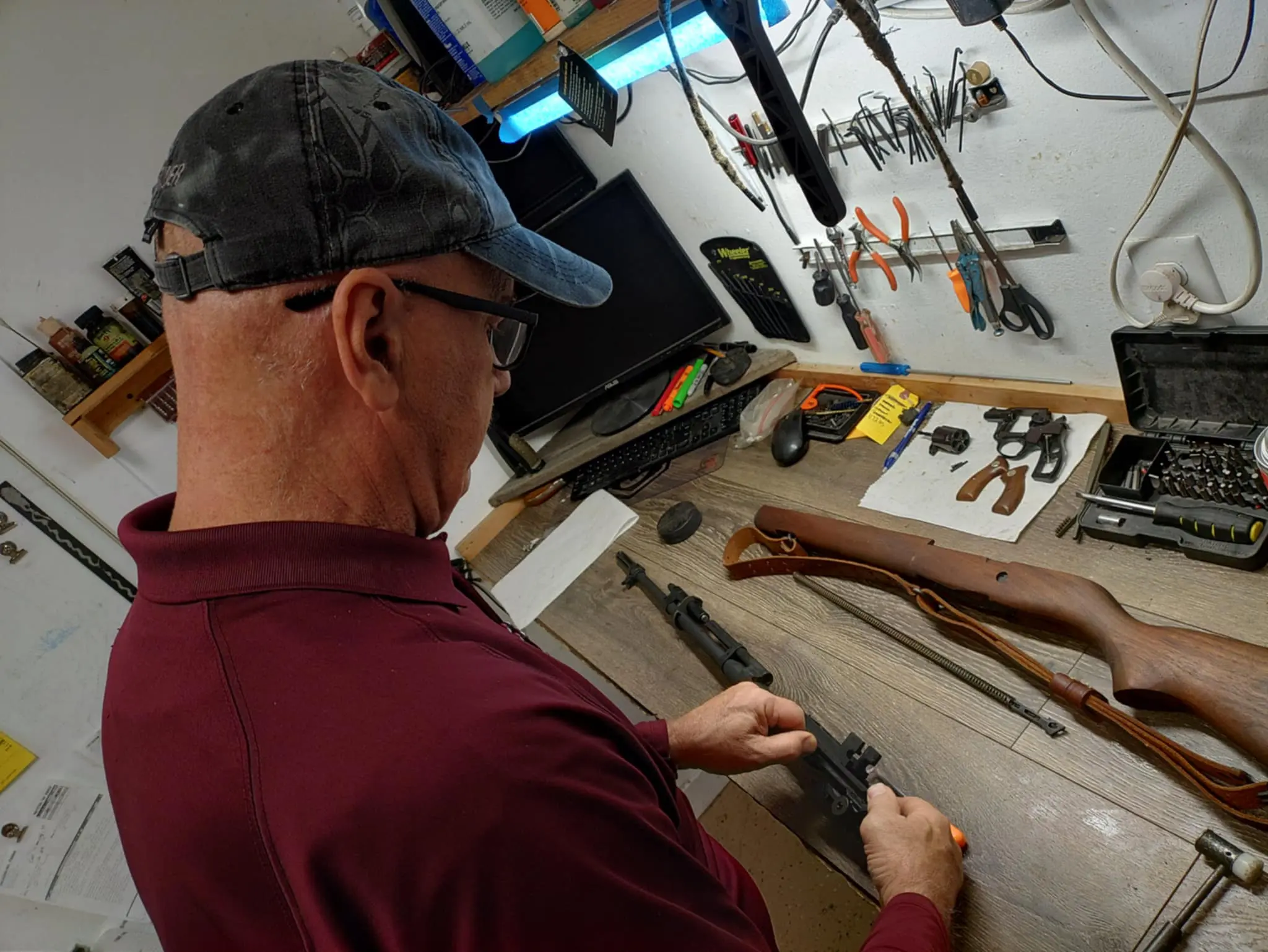An older man in a maroon shirt and baseball cap is leaning over a wooden workbench, working on a disassembled firearm, surrounded by tools and gun parts.