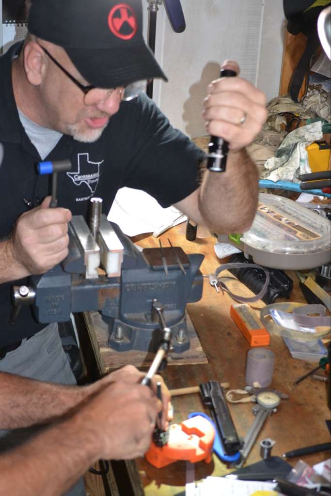 The image features a man in a black "Crosshairs Texas" shirt and baseball cap working at a messy workbench, using a vice and hand tools to perform gunsmithing or maintenance on a small part.