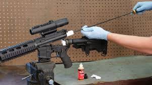 A person wearing blue gloves cleans the barrel of an assault-style rifle, which is mounted in a vice, using a cleaning rod.